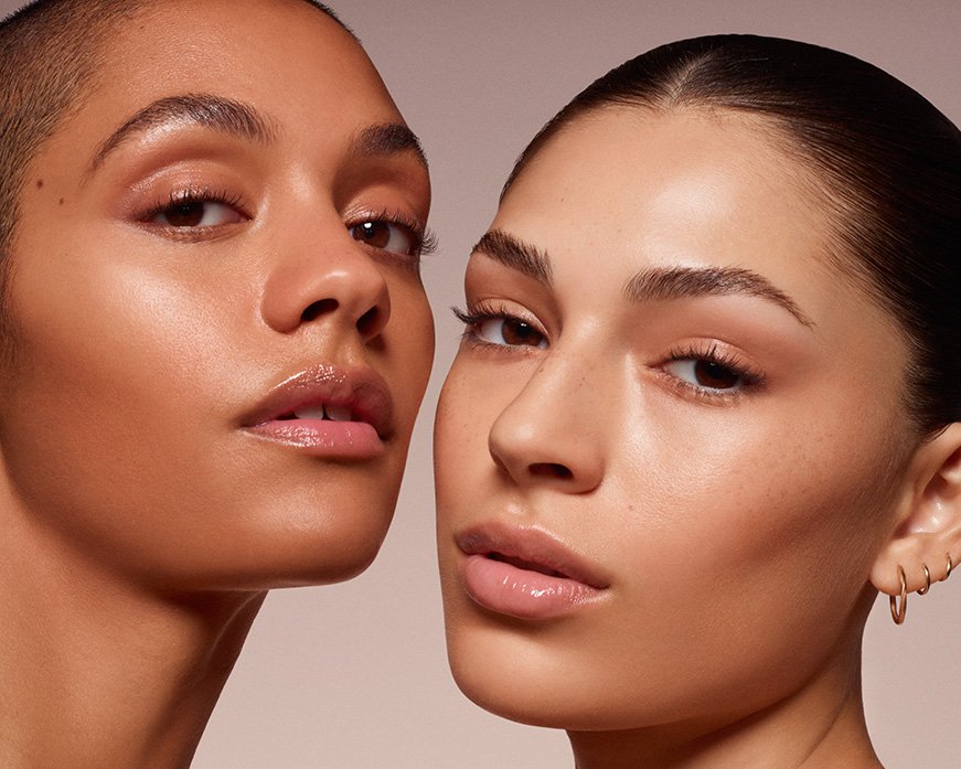 Close-up studio shot of two adult females with flawless, shiny skin, fresh makeup, and neutral expressions on a plain background.