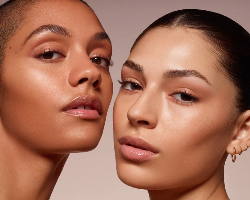 Close-up studio shot of two adult females with flawless, shiny skin, fresh makeup, and neutral expressions on a plain background.