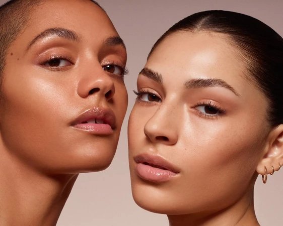 Close-up studio shot of two adult females with flawless, shiny skin, fresh makeup, and neutral expressions on a plain background.