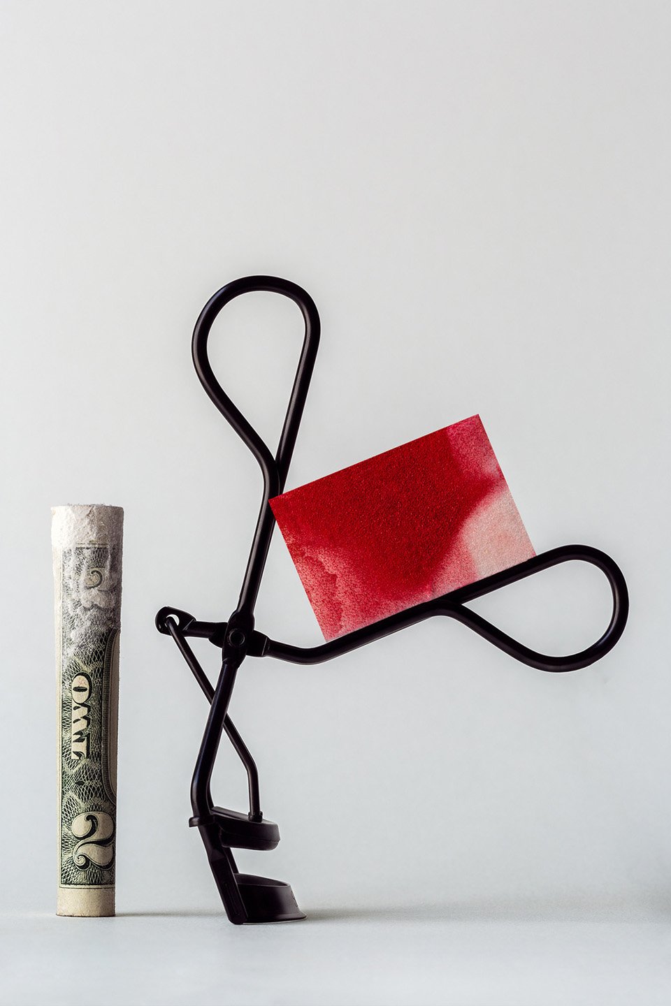 Conceptual beauty shot featuring an eyelash curler holding a makeup swatch alongside a tightly rolled two-dollar bill against a white backdrop.