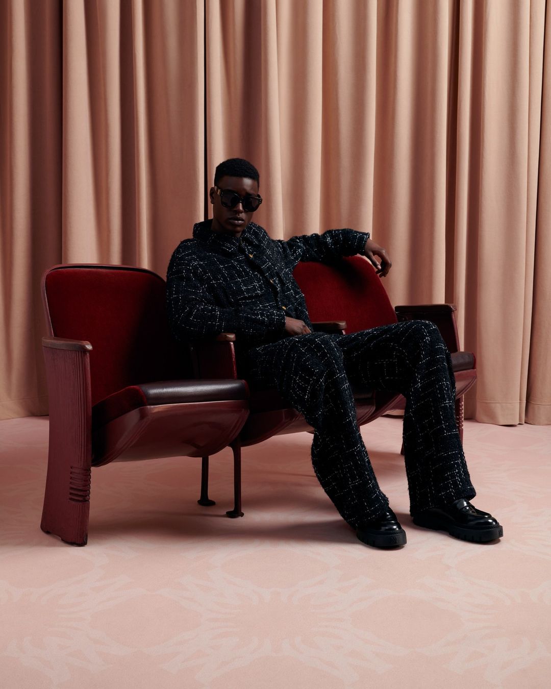 Stylish African-American man in a textured suit and sunglasses sits on a vintage red theater seat in a studio with blush drapery.