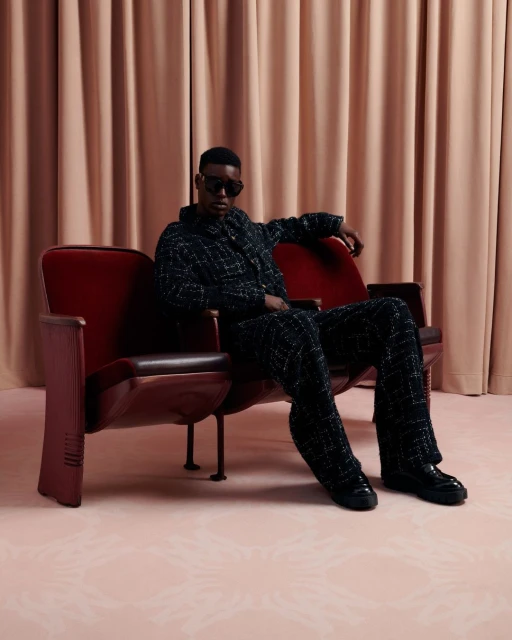 Stylish African-American man in a textured suit and sunglasses sits on a vintage red theater seat in a studio with blush drapery.