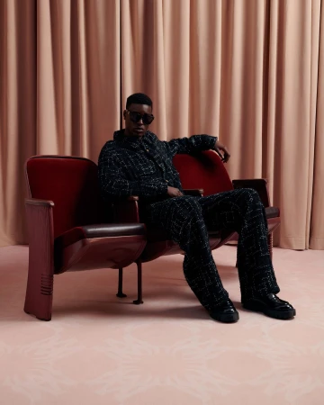 Stylish African-American man in a textured suit and sunglasses sits on a vintage red theater seat in a studio with blush drapery.