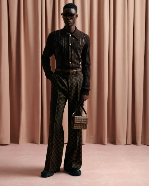 Dark-skinned male model poses in brown check pants, a brown knit shirt, sunglasses, and a small bag in a studio setting.