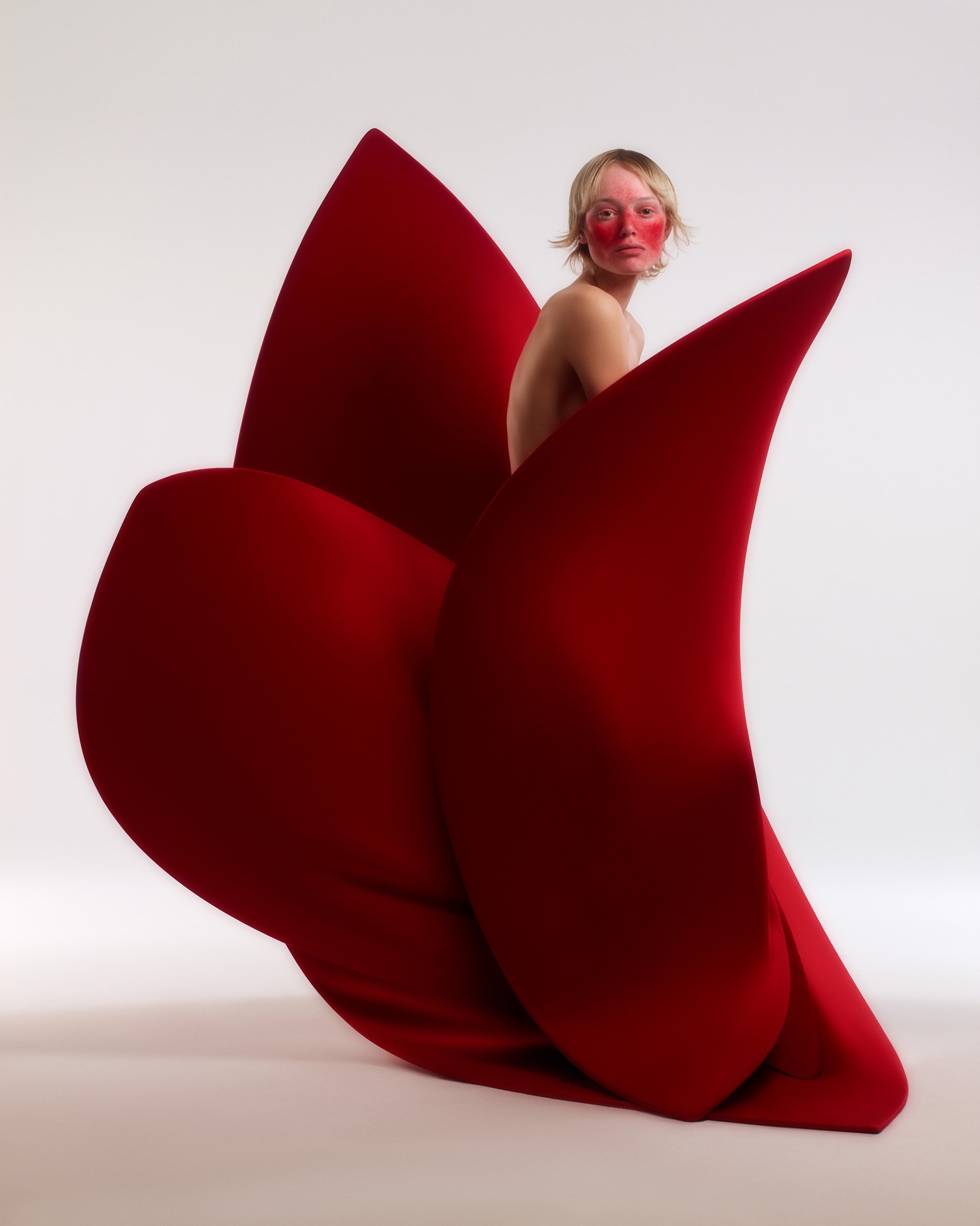 Striking image of a fair-skinned adult woman with red face paint emerging from a large, sculptural red fabric flower against a white background.