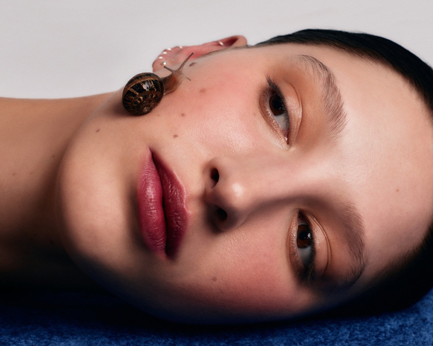 Close-up portrait of a woman with fair skin and a snail crawling on her face near her ear, set against a simple white backdrop.