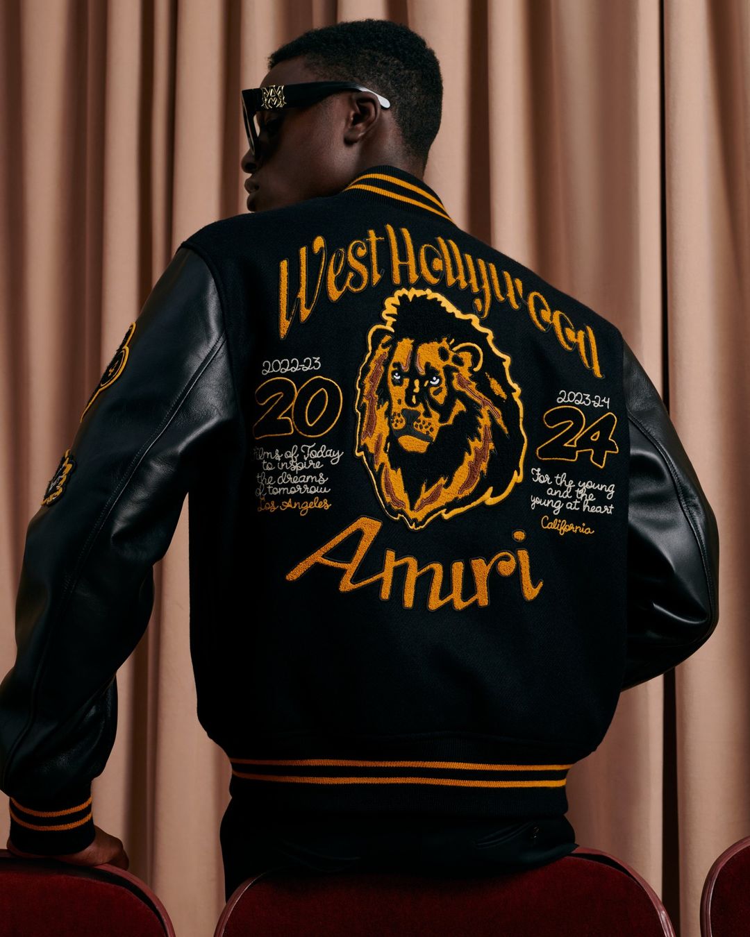 Man wearing a black and gold Amiri varsity jacket with a lion design, sunglasses, and standing in front of a beige curtain.