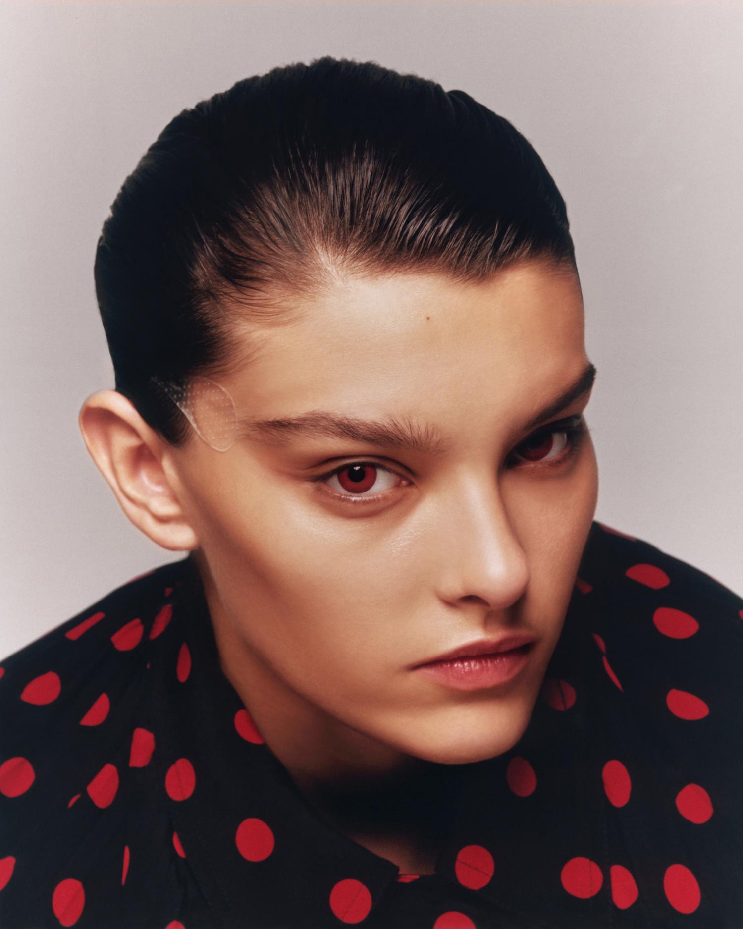Close-up portrait of a young adult female with dark hair wearing a black shirt with red polka dots, looking directly at the camera.