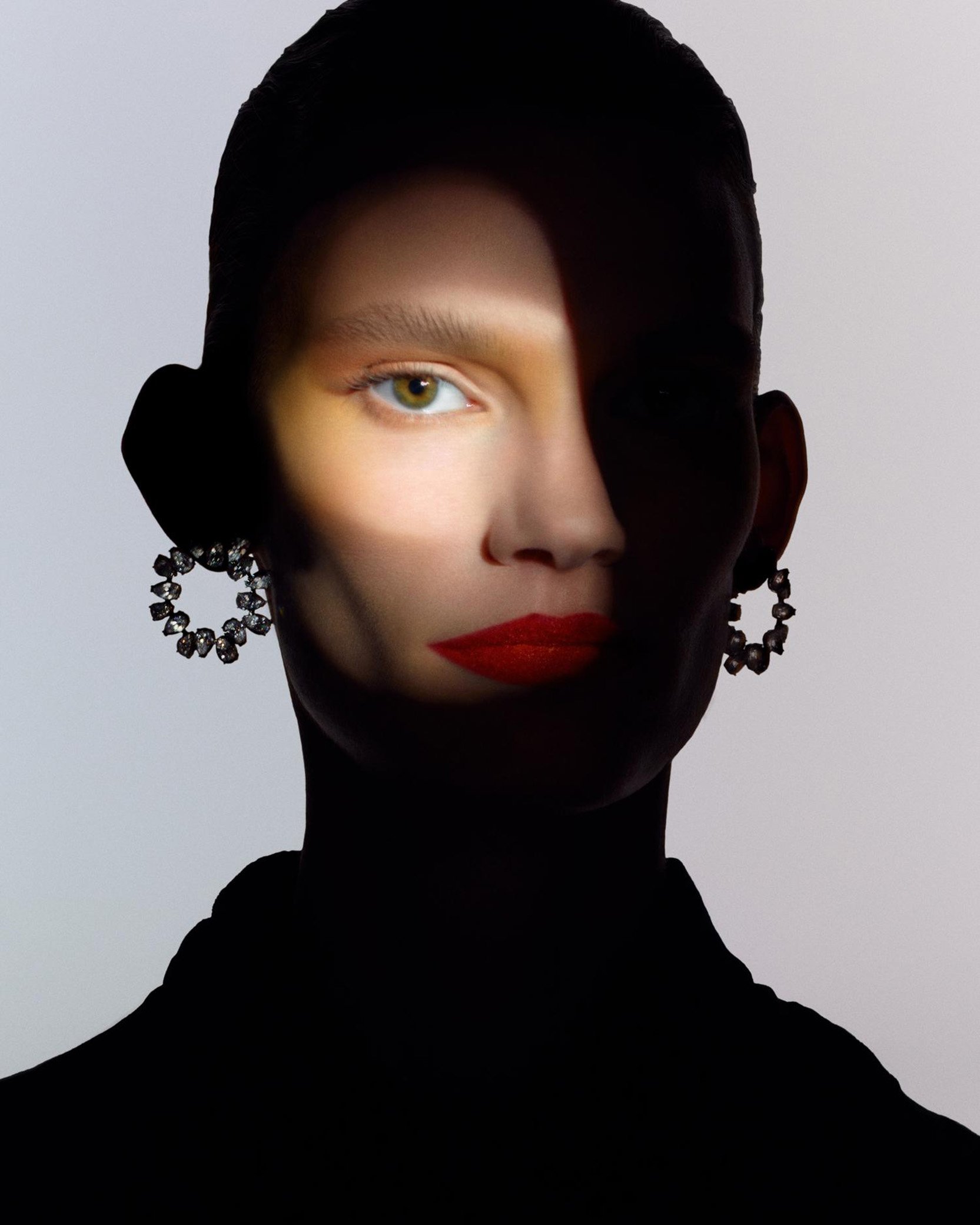 Dramatic beauty portrait of a woman with red lipstick and crystal earrings in a shadowed indoor studio, creating an alluring, high-contrast aesthetic.