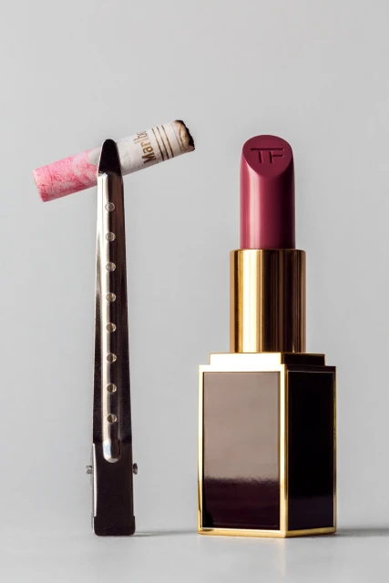 Close-up studio shot of a pink lipstick tube and a lit cigarette held by a metal clip against a neutral gray background.