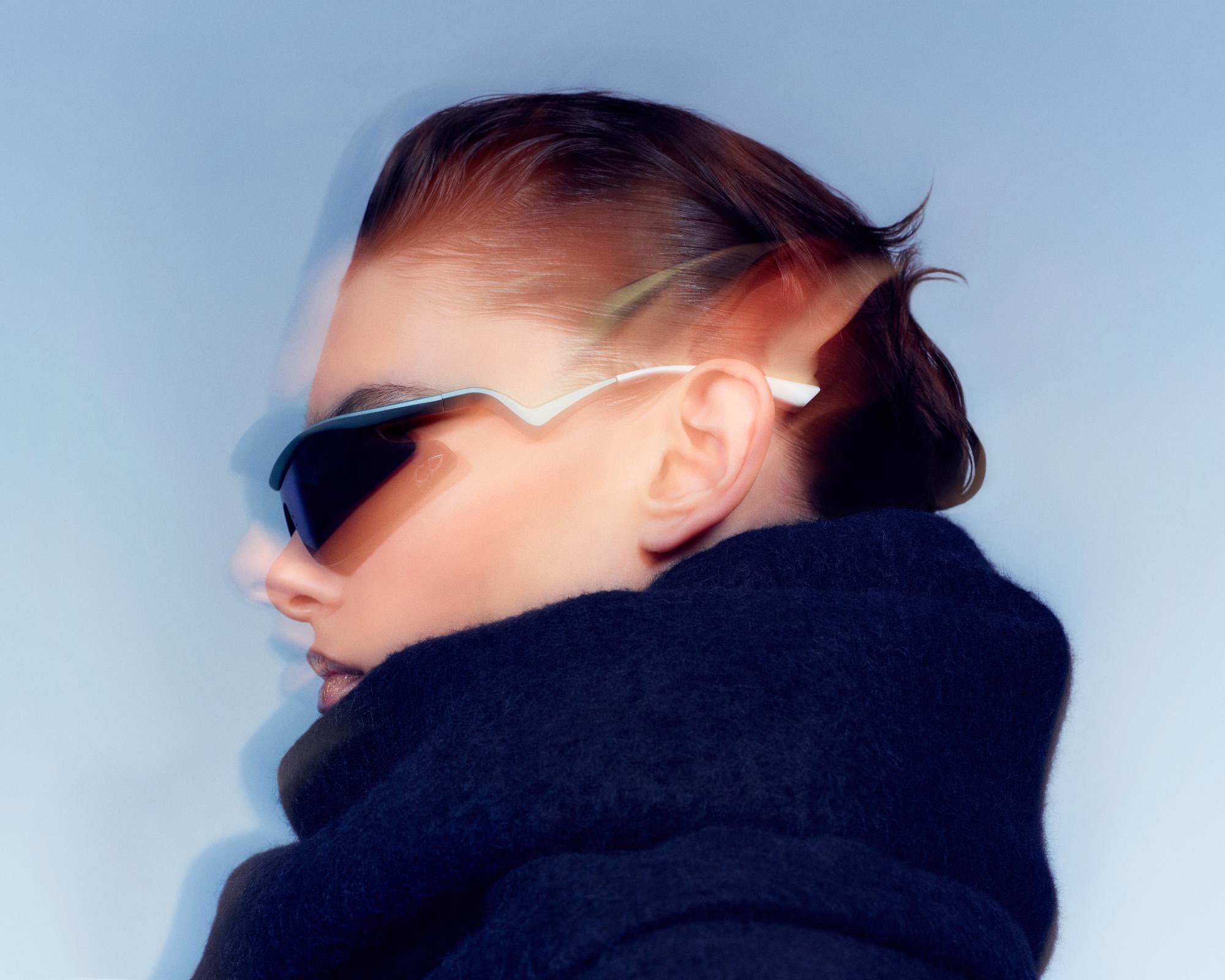 Profile portrait of a person in dark futuristic sunglasses and a heavy black scarf against a pale blue background.