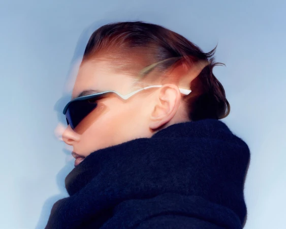 Profile portrait of a person in dark futuristic sunglasses and a heavy black scarf against a pale blue background.