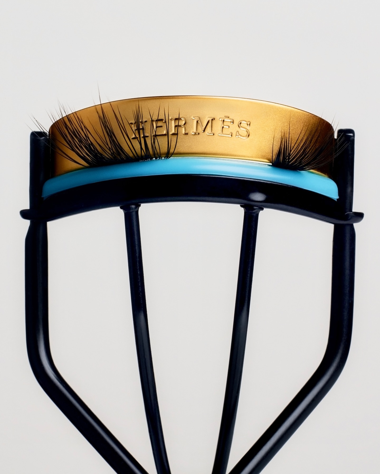 Close-up of a black and gold eyelash curler with the Hermès logo displayed on the gold handle against a white background.