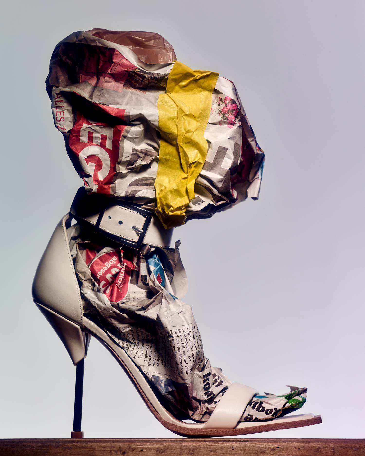 High-heeled shoe is filled with crumpled newspapers and topped with more crumpled newspapers and a yellow band on a wooden platform.