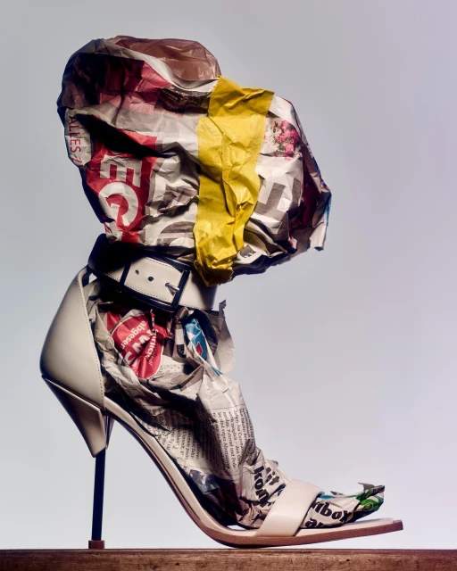 High-heeled shoe is filled with crumpled newspapers and topped with more crumpled newspapers and a yellow band on a wooden platform.