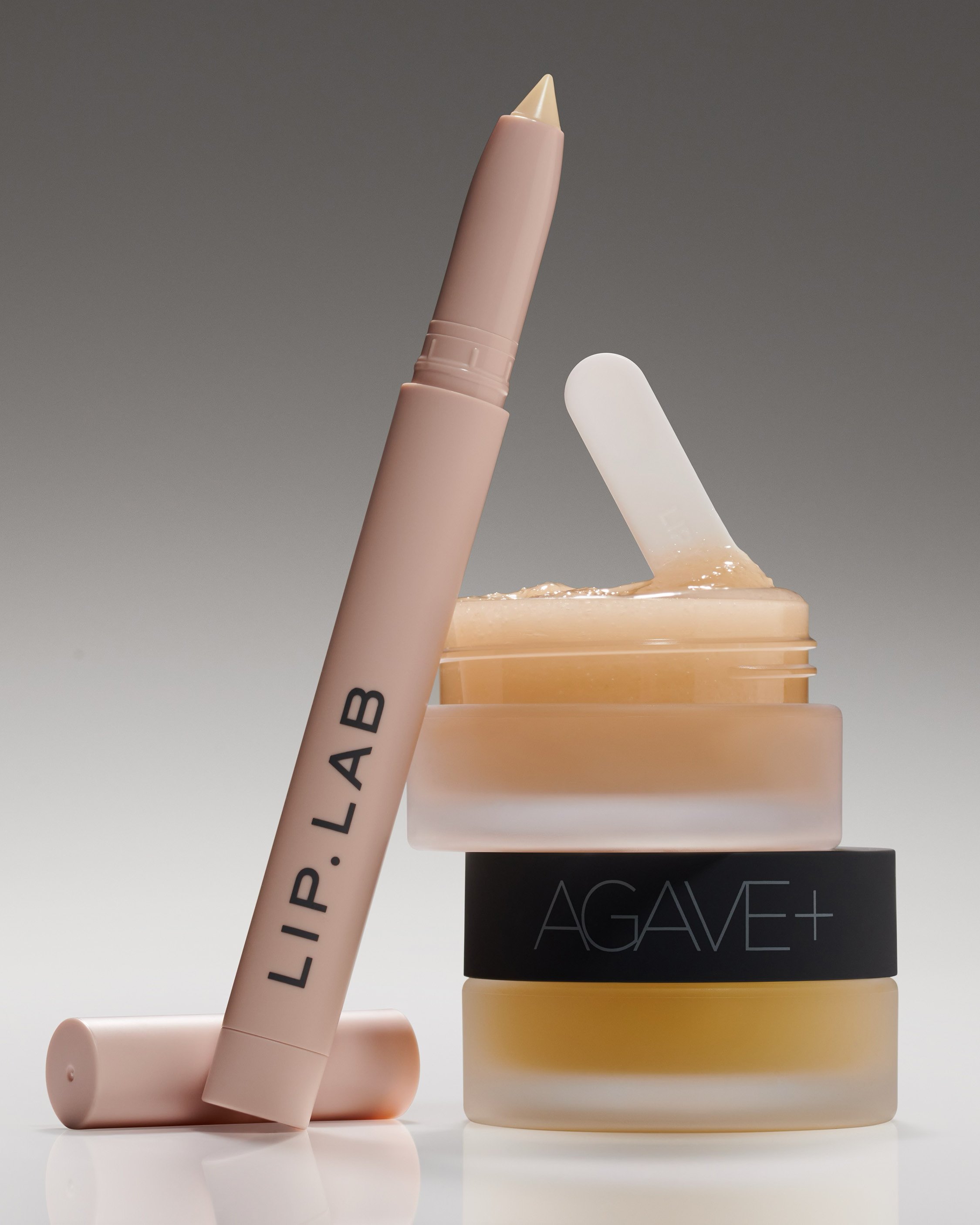 Still life shot of stacked lip care products: a lip pencil, a jar of clear balm with applicator, and a jar of golden balm.
