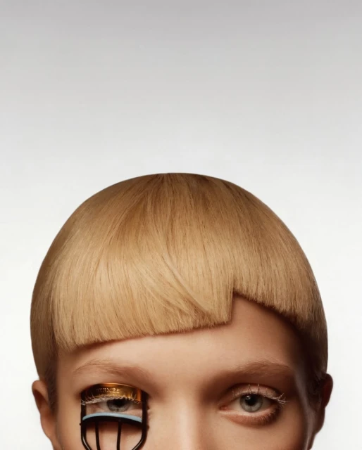 Close-up of a person with blonde hair and fair skin wearing an eyelash curler made from a bottle cap and metal on one eye.