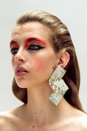 Portrait of a woman with red eyeshadow, winged eyeliner, and large metallic statement earrings against a white background