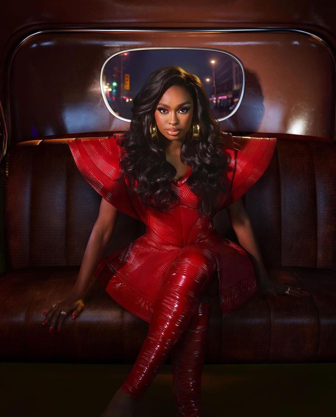 Portrait of an adult female in a red outfit sitting in a car with a night time city background. She is wearing large earrings.