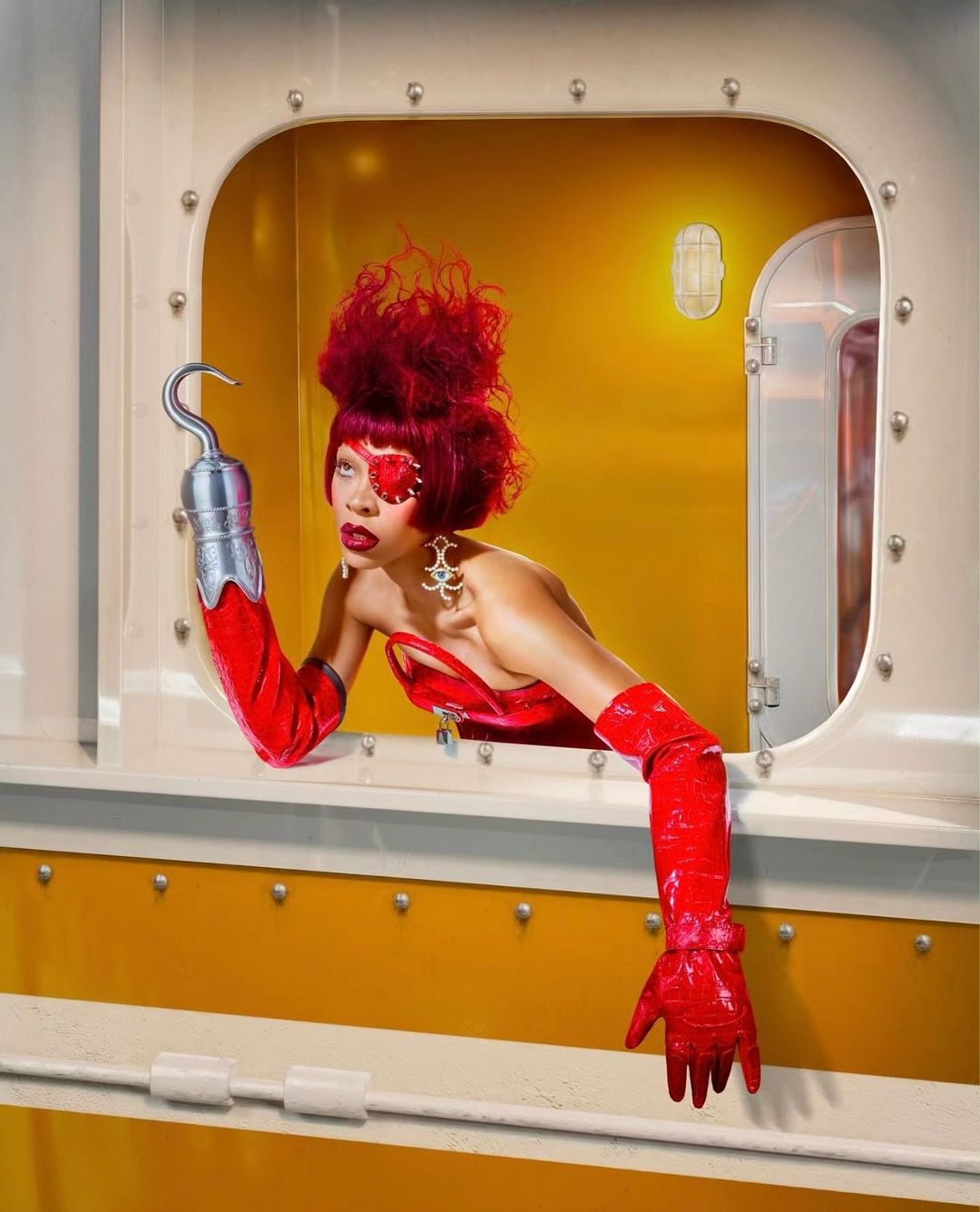 Woman with red hair and red eye makeup in yellow train car interior, wearing a red dress and hook-hand looking out of a window.