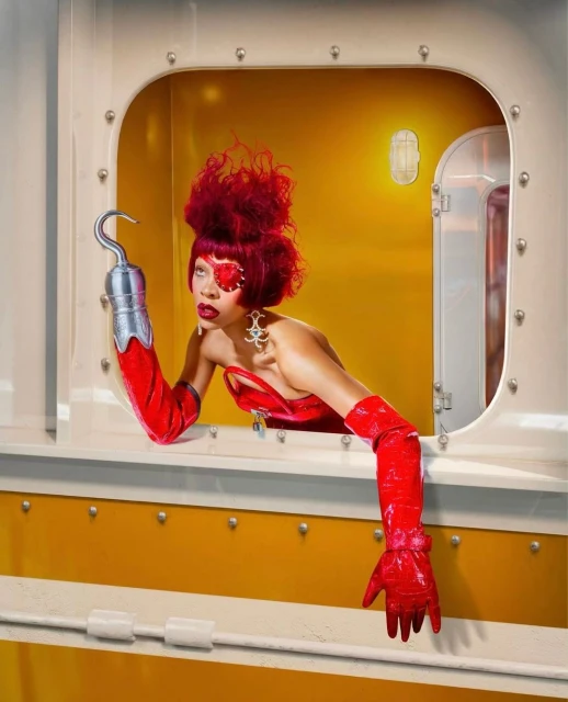 Woman with red hair and red eye makeup in yellow train car interior, wearing a red dress and hook-hand looking out of a window.