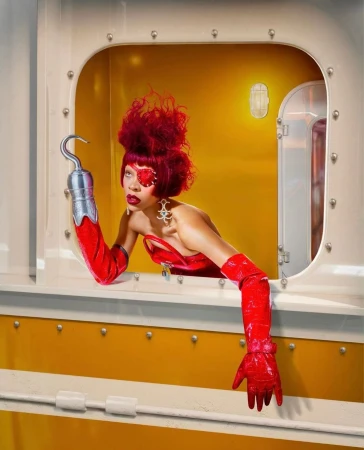 Woman with red hair and red eye makeup in yellow train car interior, wearing a red dress and hook-hand looking out of a window.