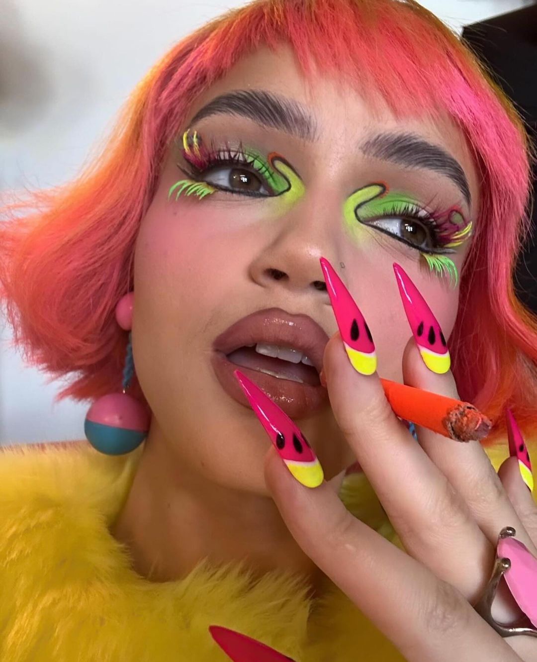 Adult woman with pink hair and colorful eyeshadow wearing earrings and holding a cigarette with watermelon-themed fingernail art.