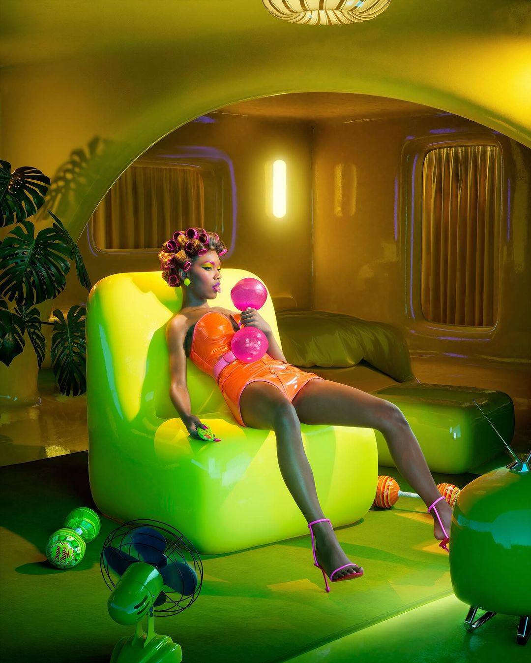 A woman in an orange outfit lounges on a neon green chair in a retro-styled room with colorful props, capturing a whimsical fashion vibe.