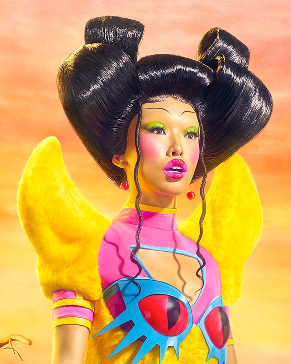 Asian woman with dramatic black sculptural updo and colorful fashion makeup poses against a blended sunset background in a portrait.
