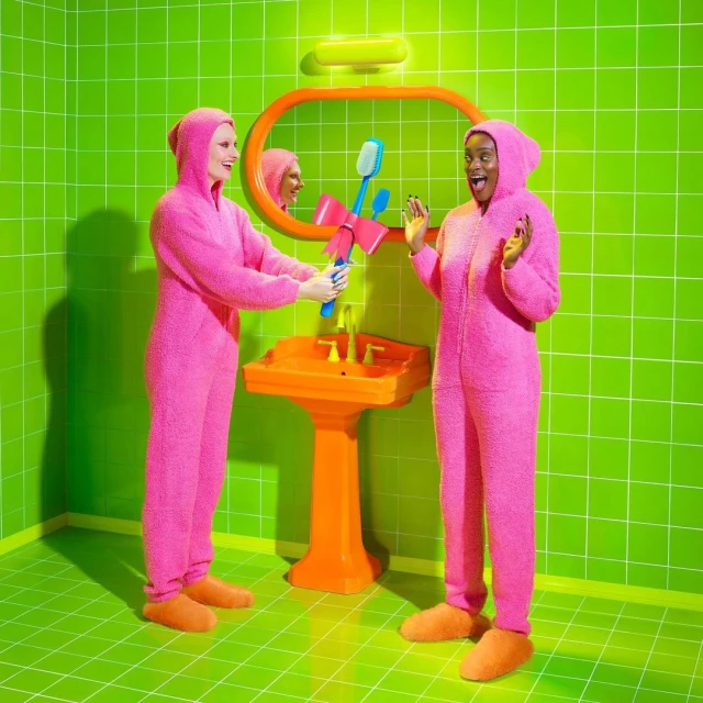 Two women in pink hooded jumpsuits smile in a green-tiled bathroom while holding a large toothbrush with a pink bow, fun and colorful.