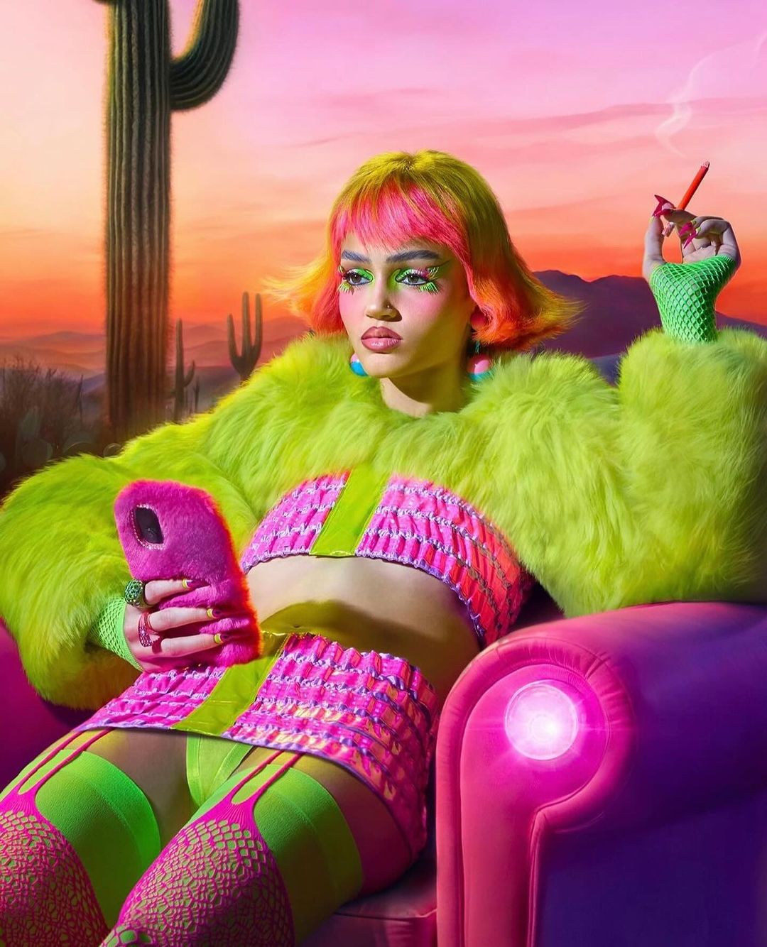 A young woman in avant-garde fashion sits in a pink chair in the desert holding a smartphone, wearing green and pink clothing and accessories.