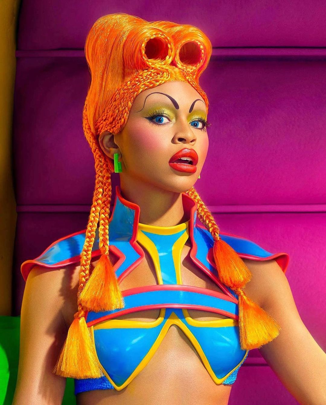 Woman with braided orange hair and bright makeup posing in a colorful outfit and background, creating a vibrant and stylized portrait.