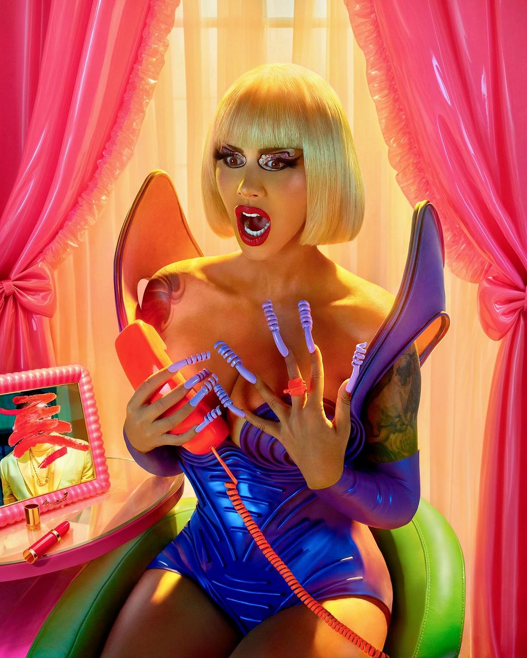 Blonde-haired woman in a blue bodysuit answers an orange landline phone at a desk with lipstick and a framed photo.