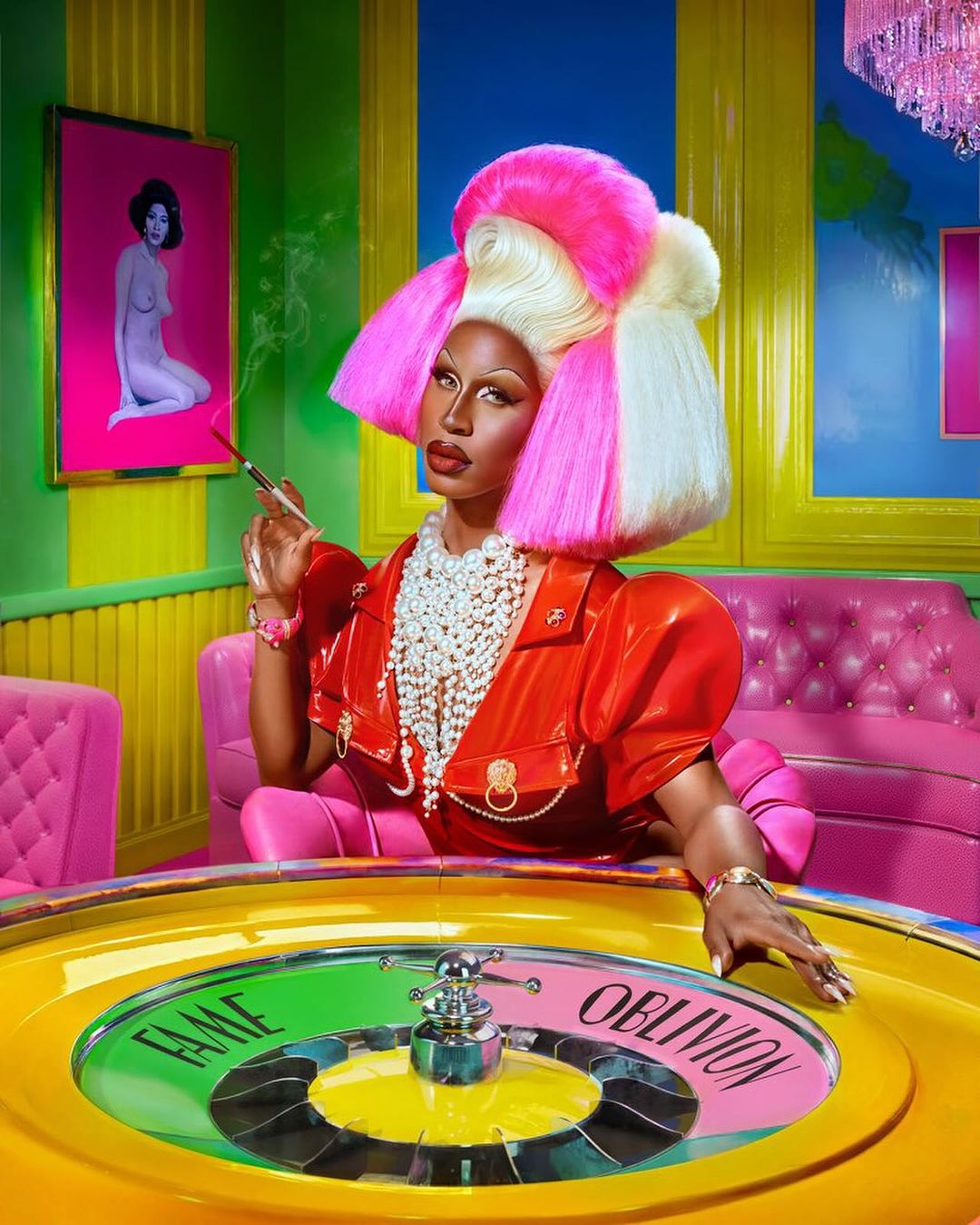 A drag queen with pink hair and an orange outfit is photographed behind a colorful roulette table in a vividly colored room.