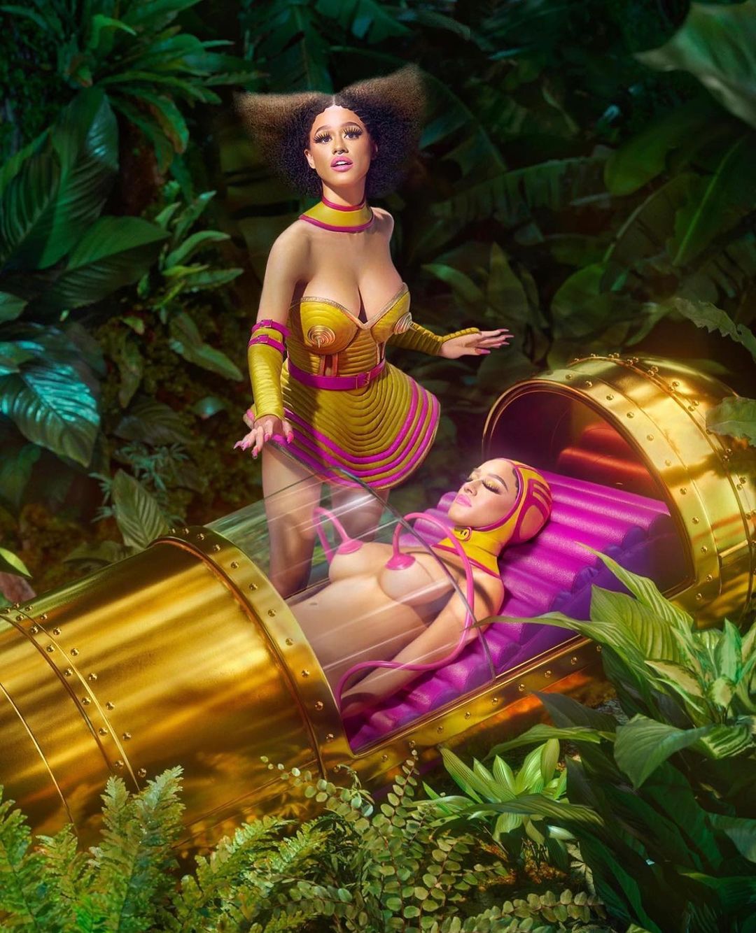Two women in fantastical costumes. One standing in a yellow dress, the other reclined inside a golden tube in a lush, green forest.