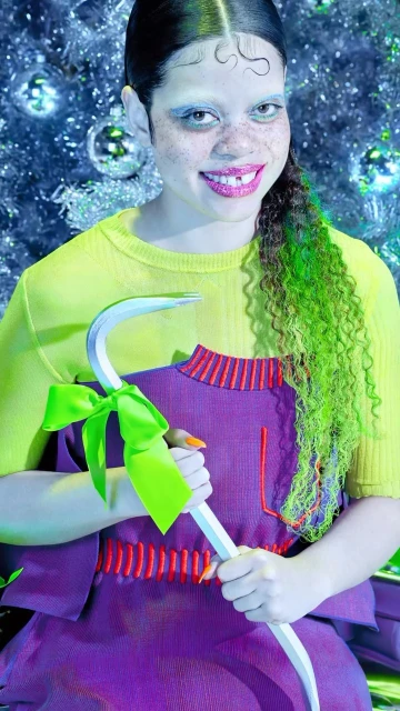 Woman in yellow top and purple dress holds a bow-tied metal bar in front of a blurred Christmas tree backdrop, showcasing a quirky, festive style.