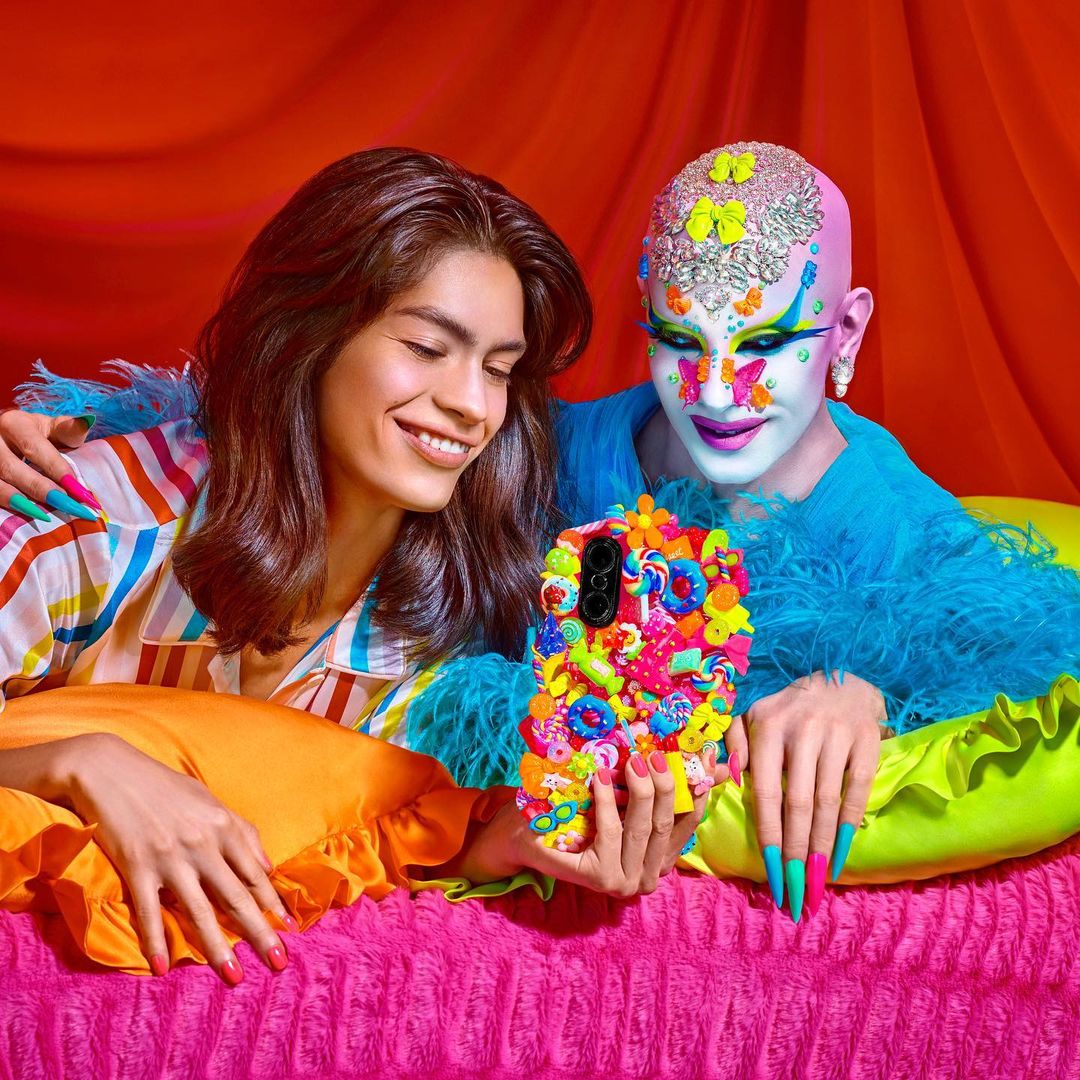 Two people pose with a smartphone in a colourful room; one has long hair and a striped shirt; the other has elaborate makeup.