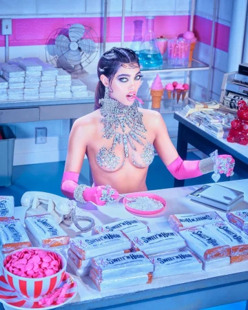 Woman in pink gloves and glittering accessories sits at a counter surrounded by candy and lab equipment in a retro-styled pink and blue setting.