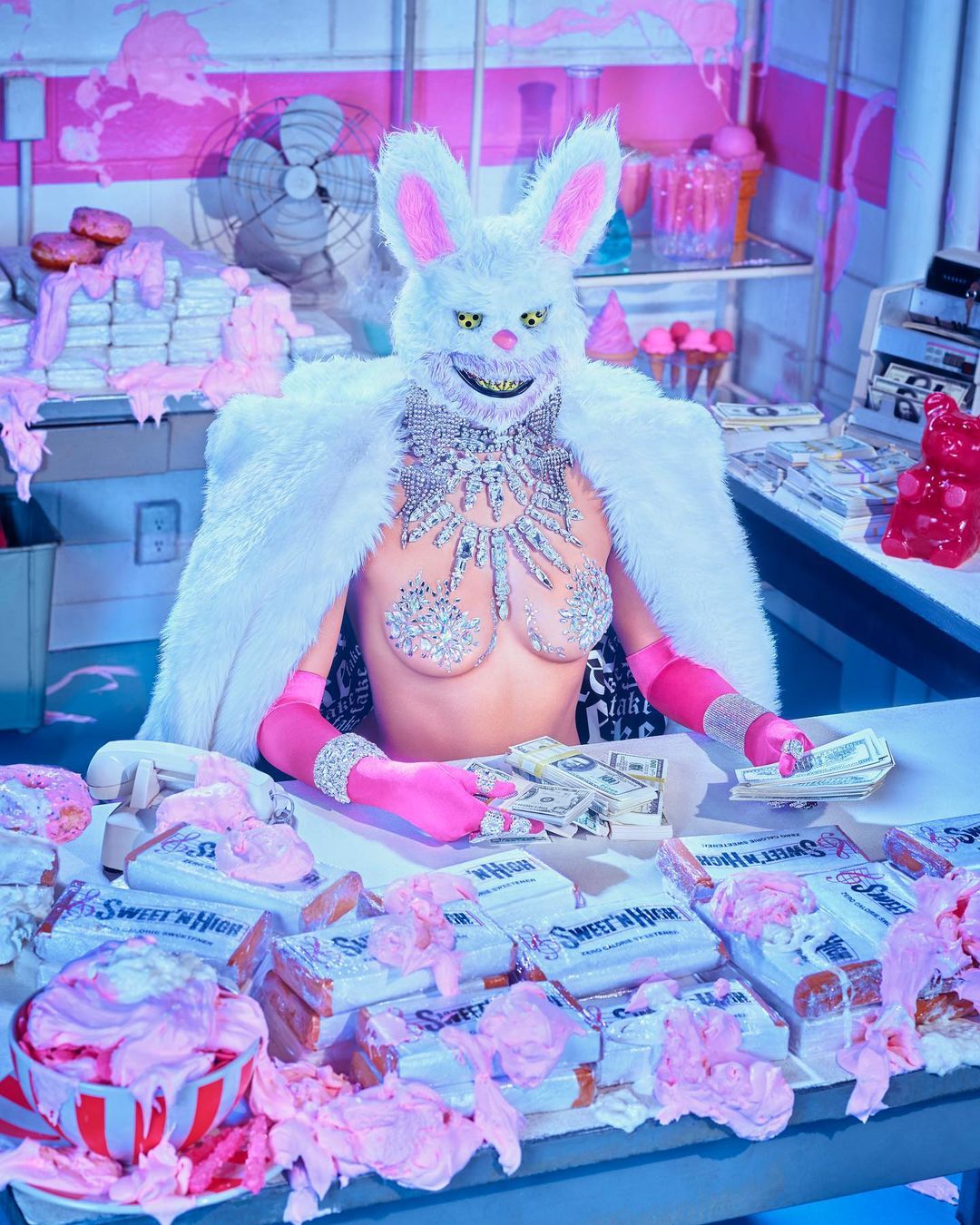 Person wearing a white bunny mask and sparkly bra stands behind a counter with snacks, money, and a vintage telephone in an indoor setting.