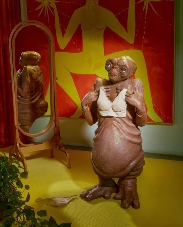 An E.T. figure stands in front of a mirror holding a bra, with a yellow and red patterned background, creating a whimsical scene.