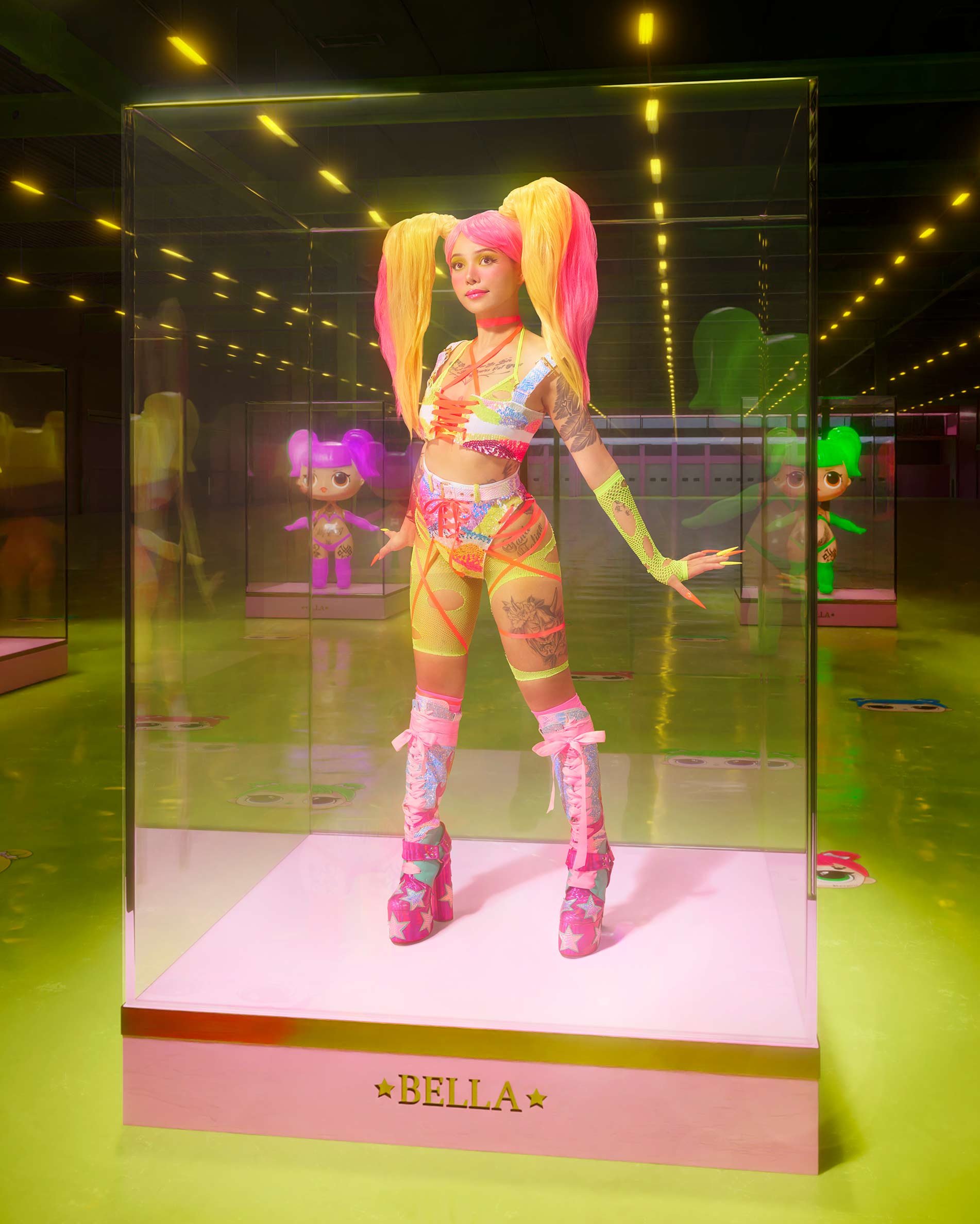 Woman with pink and yellow hair poses in a colorful outfit in a glass display case on a light green floor in a museum-like setting.