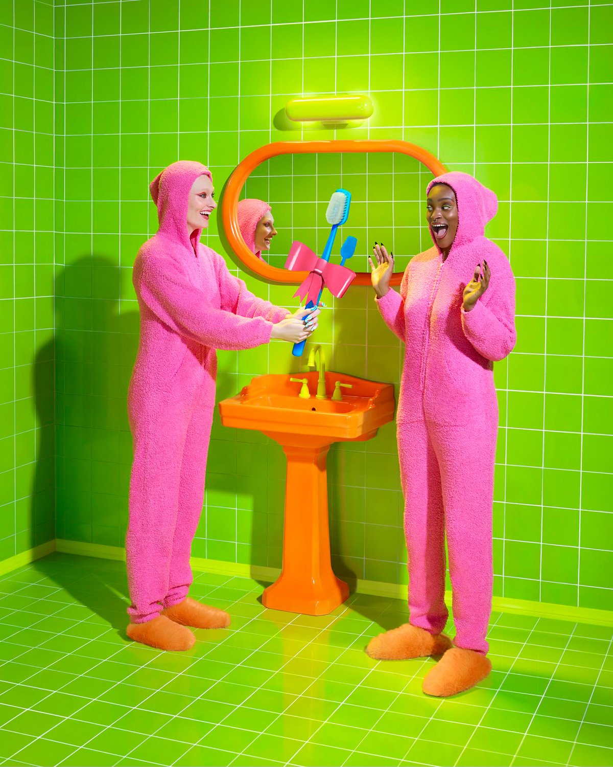 Two women in pink jumpsuits exchange toothbrush bouquet near orange sink in lime green tiled bathroom, bright and fun, studio shot.