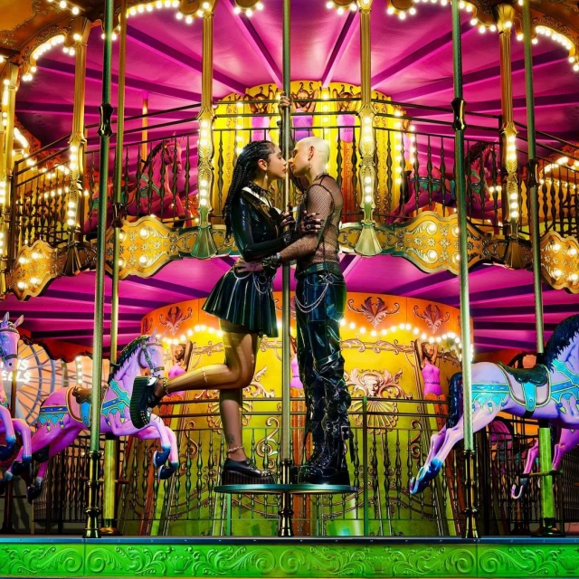 A couple embraces on a brightly lit carousel with colorful horses; the woman wears a black outfit, and the man wears a black mesh top.