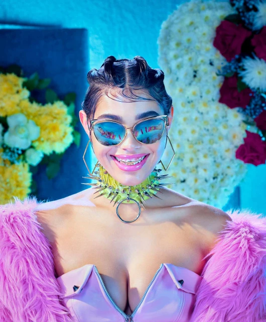 Close-up of a smiling adult woman with braces, sunglasses, spike choker, and a pink fur stole against a floral backdrop and blue wall.