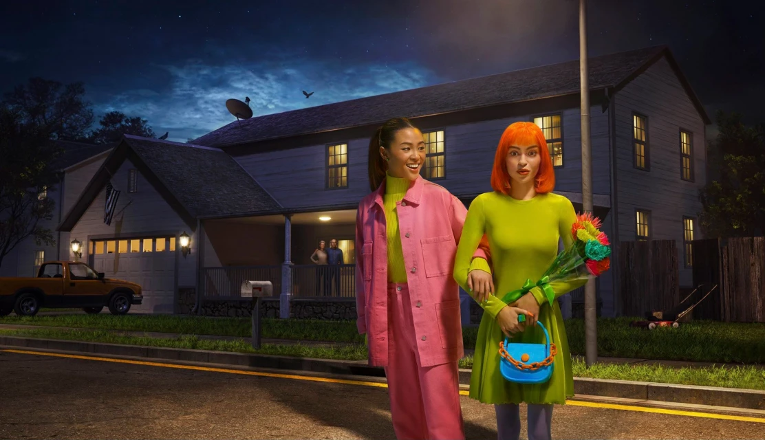 Two women in colorful outfits walk on a street in a residential neighborhood at night; a house and truck are in the background.