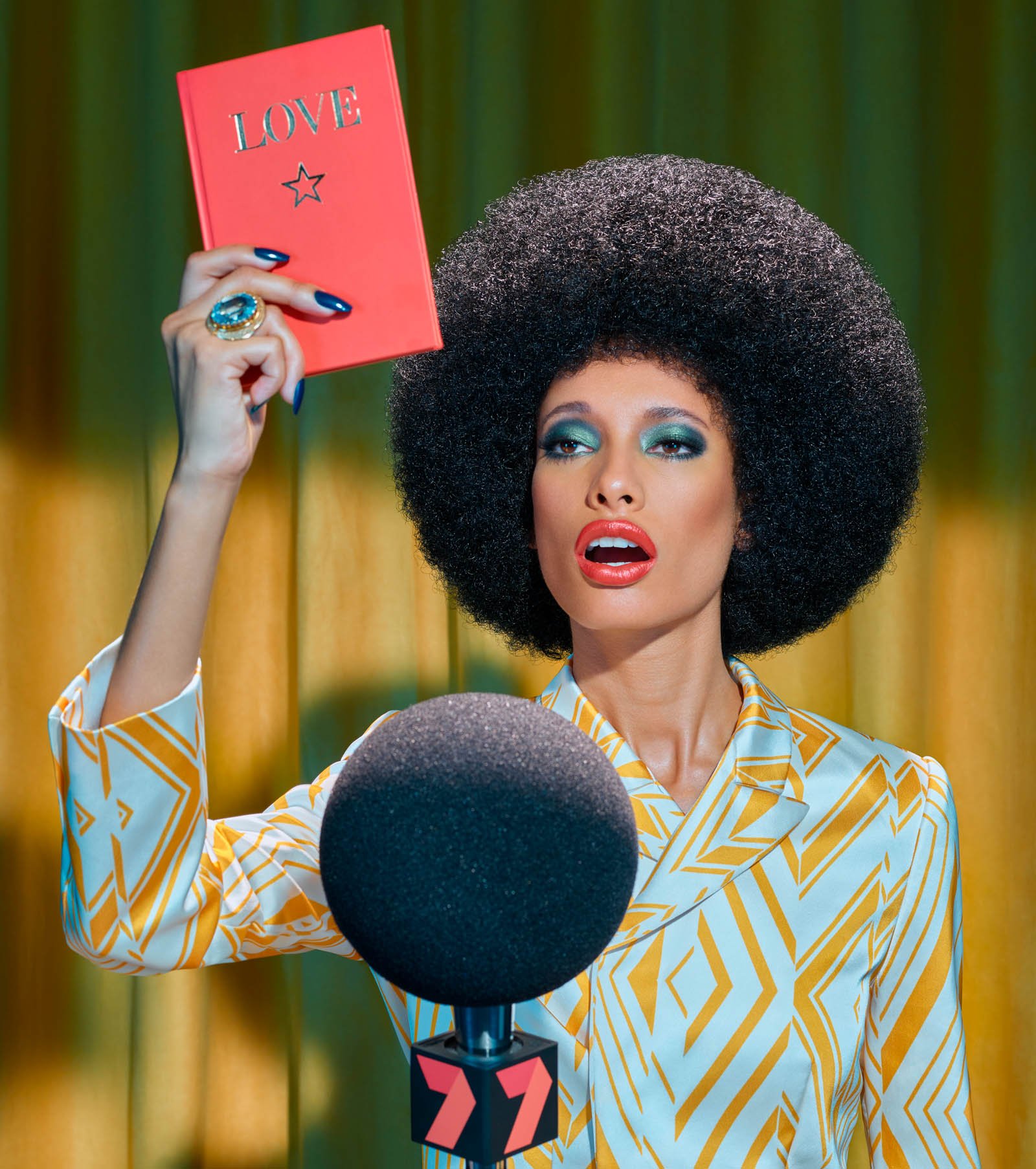 Stylish woman with a large afro stands in front of a microphone holding a red book with 'LOVE' in gold text.