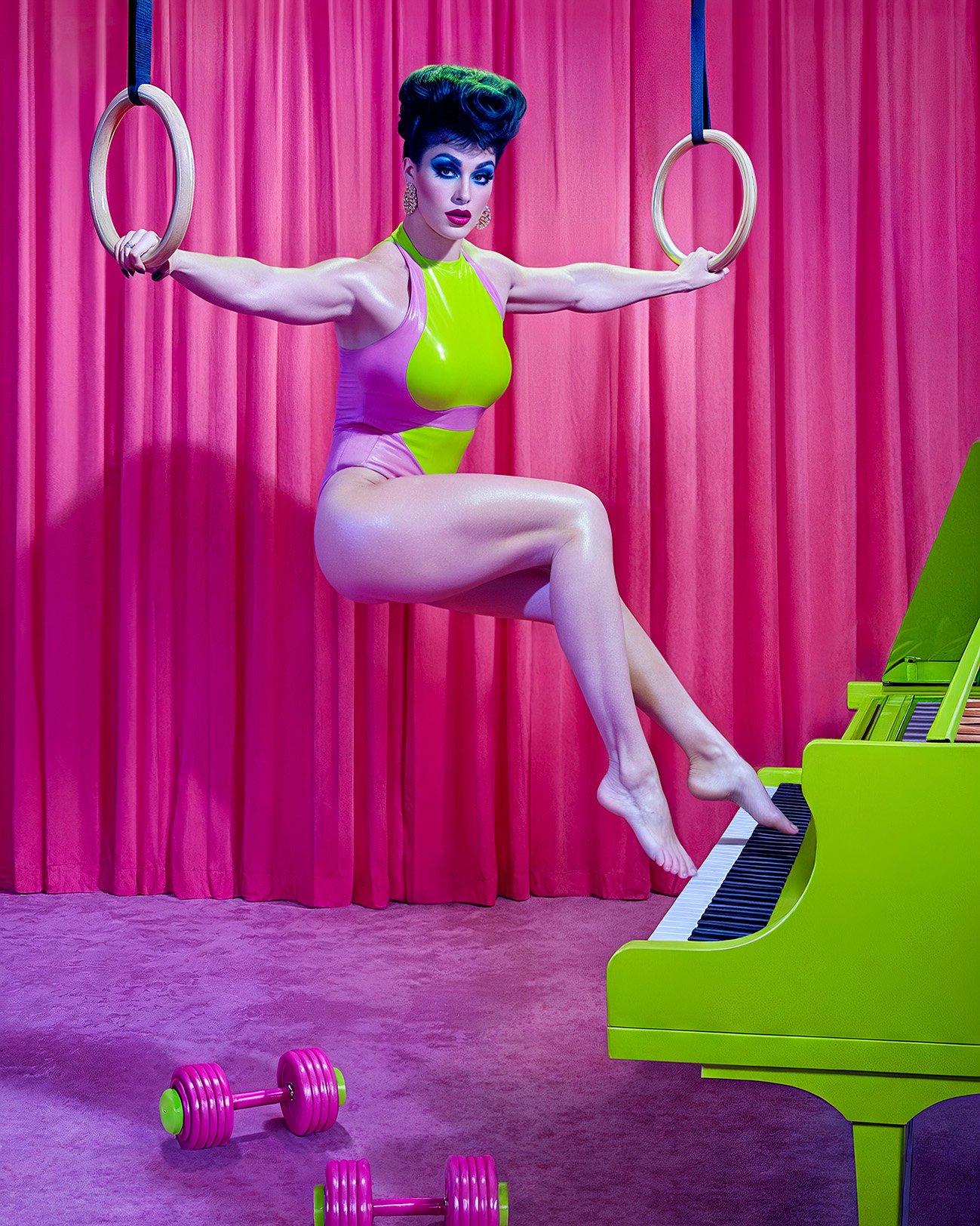 A woman in a pink and green leotard balances on gymnastics rings while touching a lime green piano with her toes in studio shot.