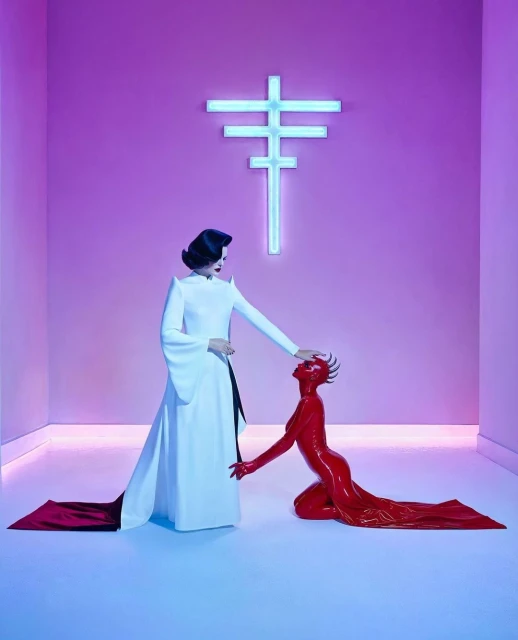 Artistic studio shot of a woman in white standing next to a kneeling woman in red latex, with a neon cross in the background.