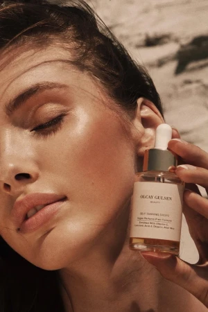 Close-up shot of a woman holding an Olcay Gulsen Beauty serum bottle, showcasing skincare and beauty in a sunlit outdoor setting.