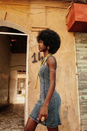 Black woman with afro wearing a denim sleeveless dress standing beside a weathered textured wall